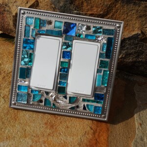 May include: A double light switch cover with a mosaic design featuring blue and green glass tiles. The cover has a silver metal frame with a decorative border. A mermaid design is visible in the centre of the mosaic.