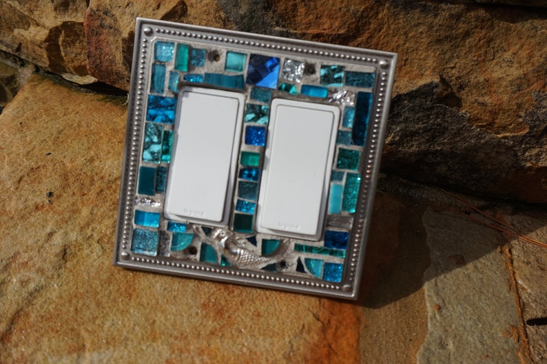 May include: A double light switch cover with a mosaic design featuring blue and green glass tiles. The cover has a silver metal frame with a decorative border. A mermaid design is visible in the centre of the mosaic.