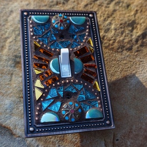 Pu&ograve; includere: Una placca per interruttore della luce decorativa con un design a mosaico con piastrelle di vetro turchese, blu, oro e arancio. La placca dell'interruttore ha una cornice color rame con un bordo perlato.