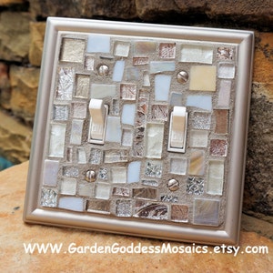 May include: A silver light switch cover with a mosaic design in shades of white, gray, and silver. The mosaic is made of small, irregular pieces of glass and tile. The switch cover has two toggle switches with the word "ON" printed on them.