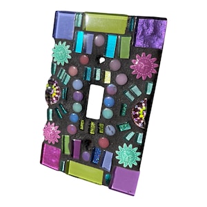 May include: A colorful mosaic light switch cover with a black background. The cover features a variety of shapes and colors, including green, blue, purple, and pink. There are also small sun-shaped designs throughout the cover.