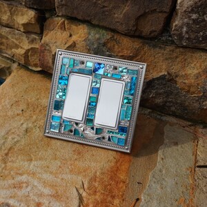 May include: Double light switch cover with a mosaic design featuring blue and turquoise glass tiles. The cover has a silver metal frame.