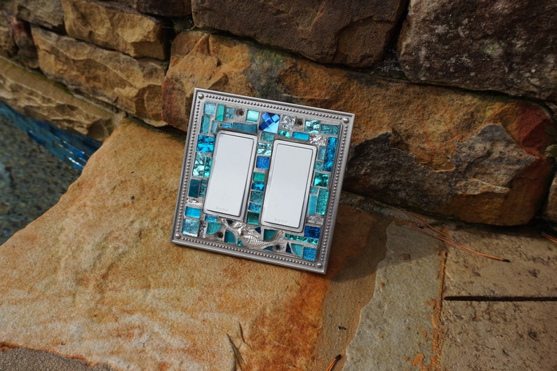 May include: Double light switch cover with a mosaic design featuring blue and turquoise glass tiles. The cover has a silver metal frame.