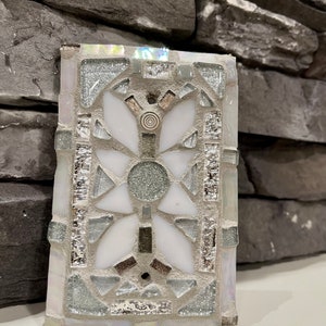 Puede incluir: Un azulejo de mosaico blanco con un dise&ntilde;o geom&eacute;trico plateado y gris. El azulejo tiene un patr&oacute;n floral en el centro.