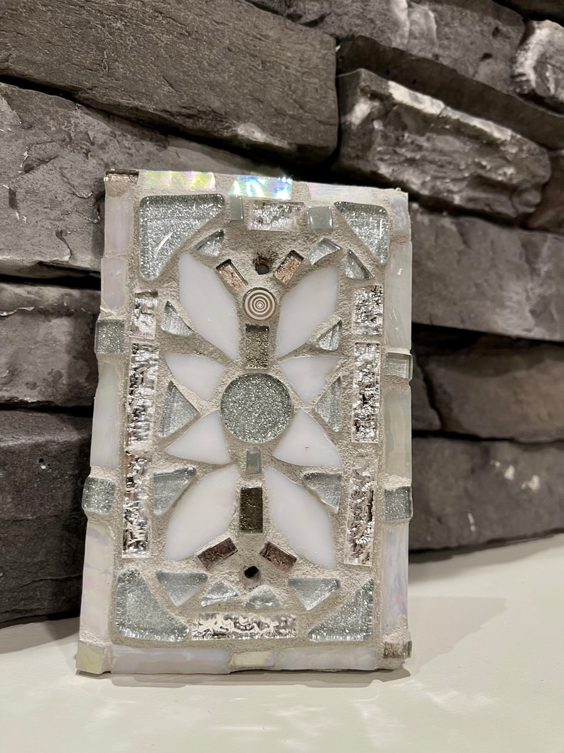 Puede incluir: Un azulejo de mosaico blanco con un dise&ntilde;o geom&eacute;trico plateado y gris. El azulejo tiene un patr&oacute;n floral en el centro.