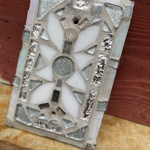 Puede incluir: Un azulejo de mosaico blanco y plateado con un dise&ntilde;o floral. El azulejo est&aacute; hecho de peque&ntilde;os trozos de vidrio y est&aacute; colocado en un mortero gris.