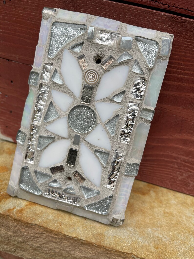 Puede incluir: Un azulejo de mosaico blanco y plateado con un dise&ntilde;o floral. El azulejo est&aacute; hecho de peque&ntilde;os trozos de vidrio y est&aacute; colocado en un mortero gris.