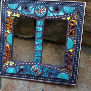 Pu&ograve; includere: Una placca di copertura per interruttore doppio con un design a mosaico. Il mosaico presenta piastrelle di vetro turchese, blu, oro e arancio. La copertura &egrave; in rame e ha una finitura anticata.