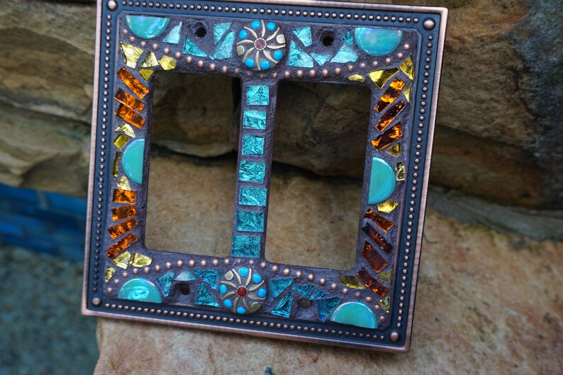 Pu&ograve; includere: Una placca di copertura per interruttore doppio con un design a mosaico. Il mosaico presenta piastrelle di vetro turchese, blu, oro e arancio. La copertura &egrave; in rame e ha una finitura anticata.