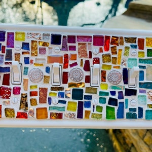 May include: A white light switch cover with a colorful mosaic design. The mosaic is made of small pieces of glass in various colors, including red, yellow, blue, green, and purple. The switch cover has three toggle switches.