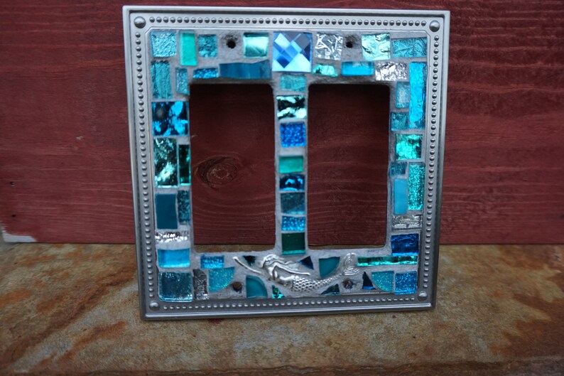 May include: A silver double light switch cover with a mosaic design featuring blue and turquoise glass tiles. A mermaid is featured in the centre of the design.