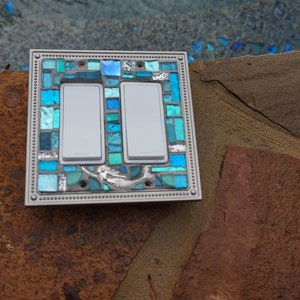 May include: A double light switch cover featuring a mermaid design. The cover is made of mosaic tiles in shades of blue and green.