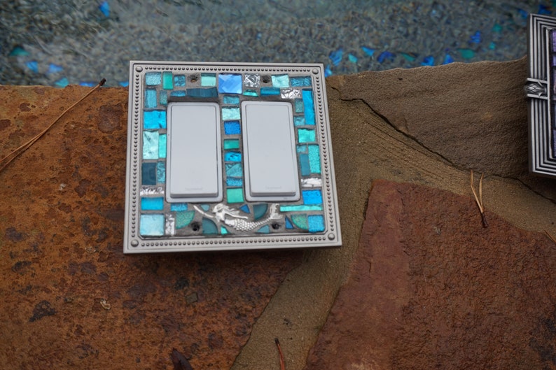 May include: A double light switch cover featuring a mermaid design. The cover is made of mosaic tiles in shades of blue and green.
