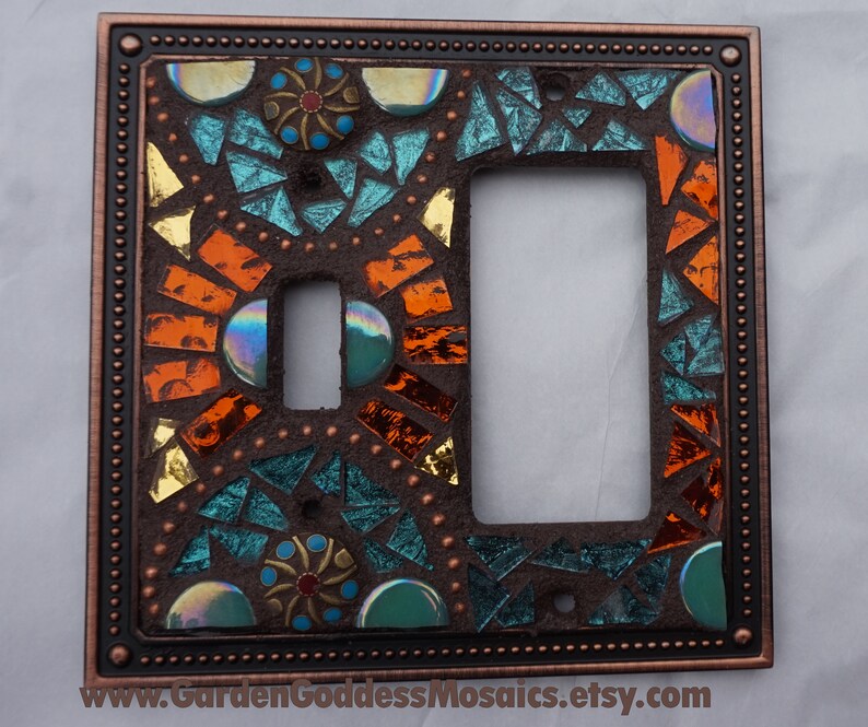 Pu&ograve; includere: Una placca per interruttore della luce color rame con un design a mosaico con piastrelle di vetro turchese, arancio e oro. Il design del mosaico &egrave; intricato e presenta forme geometriche e motivi.