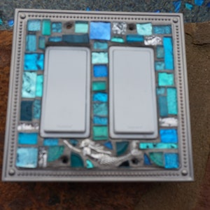 May include: A double light switch cover featuring a mermaid design. The cover is crafted from mosaic tiles in shades of blue and green.