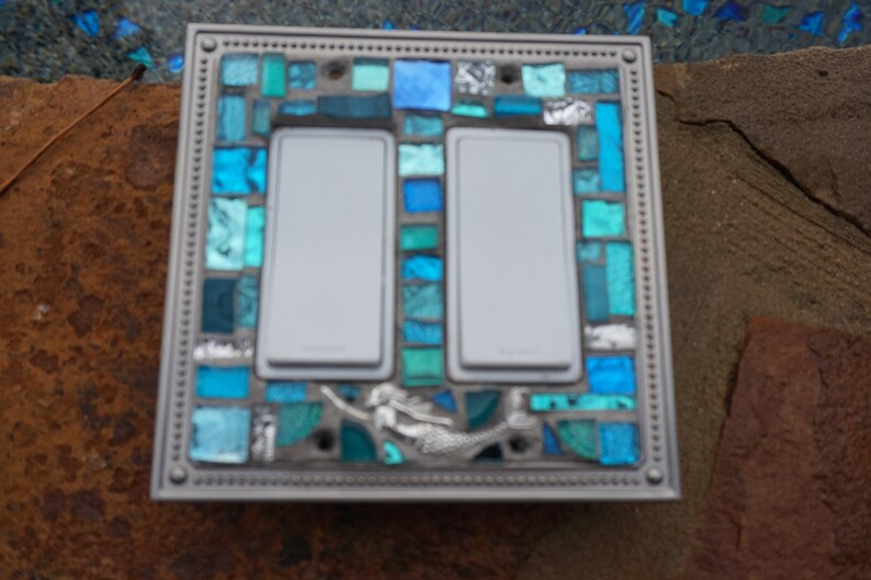 May include: A double light switch cover featuring a mermaid design. The cover is crafted from mosaic tiles in shades of blue and green.