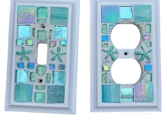 May include: Two white light switch and outlet covers with a mosaic design. The mosaic is made of small, iridescent blue and green tiles. The covers feature a starfish design.