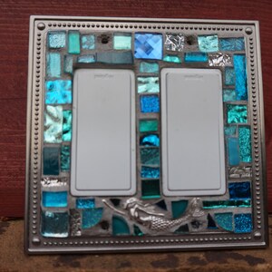May include: A double light switch cover with a mosaic design featuring blue and green glass tiles. A mermaid is depicted in the centre of the cover.