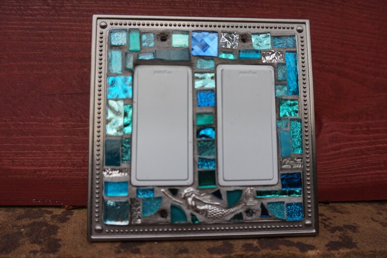 May include: A double light switch cover with a mosaic design featuring blue and green glass tiles. A mermaid is depicted in the centre of the cover.