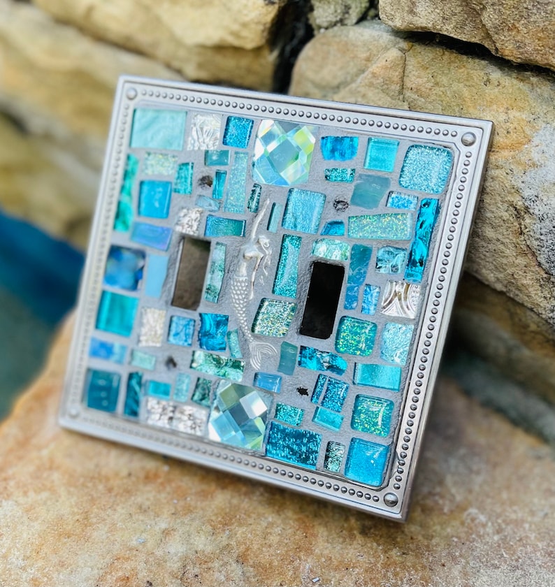 May include: A light switch cover with a mosaic design featuring blue and green glass tiles, a mermaid, and a crystal accent. The cover is made of metal and has a silver finish.