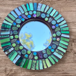 May include: A round mirror with a green mosaic frame. The frame is made of small, green glass tiles arranged in a circular pattern. The mirror reflects a blue sky with white clouds.
