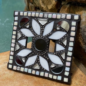 May include: A black and white mosaic light switch cover with a floral design. The cover is made of small, iridescent tiles and features a mirrored center.