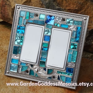 May include: A silver light switch cover with a mosaic of blue and green glass tiles. The mosaic features a mermaid design and the text "Legrand" is visible on the switch plates.