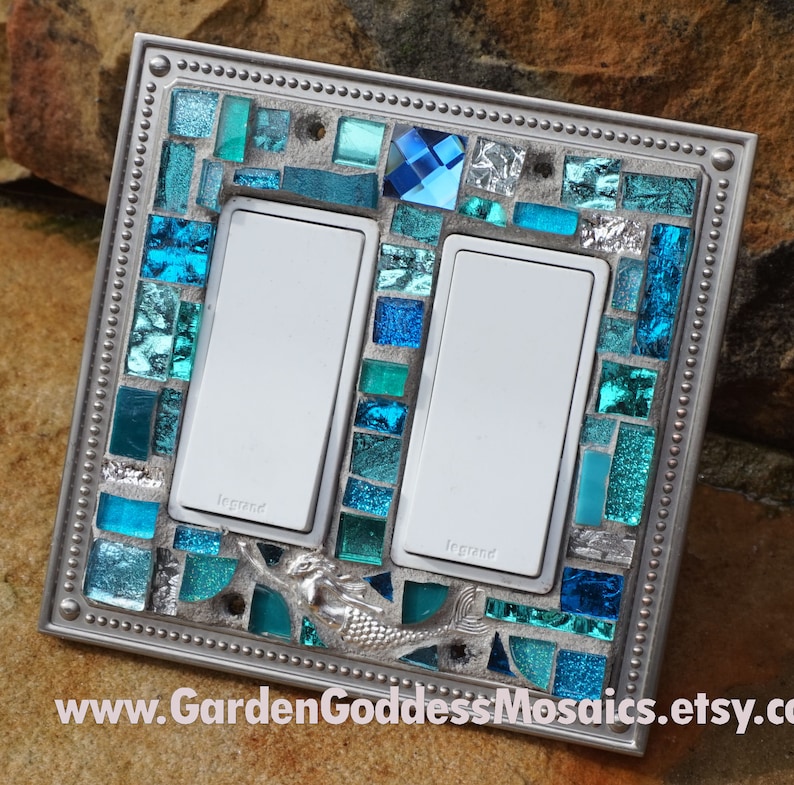 May include: A silver light switch cover with a mosaic of blue and green glass tiles. The mosaic features a mermaid design and the text "Legrand" is visible on the switch plates.