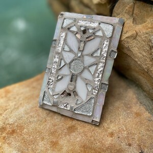 Puede incluir: Un azulejo de mosaico blanco y plateado con un dise&ntilde;o floral. El azulejo est&aacute; hecho de peque&ntilde;os trozos de vidrio y est&aacute; colocado en un mortero blanco.