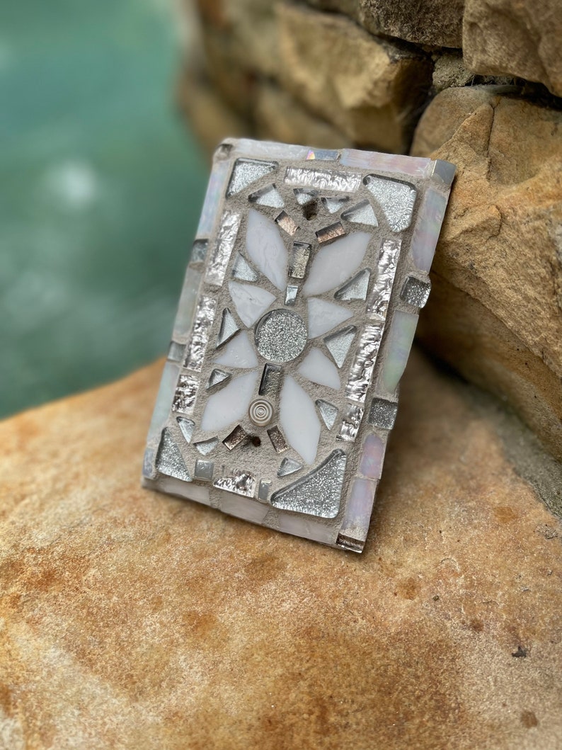 Puede incluir: Un azulejo de mosaico blanco y plateado con un dise&ntilde;o floral. El azulejo est&aacute; hecho de peque&ntilde;os trozos de vidrio y est&aacute; colocado en un mortero blanco.