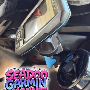 May include: A black and gray Garmin GPS device mounted on a black and blue bracket. The bracket is attached to a black surface. The text "SEADOO GARMIN MOUNT" is displayed in a colorful, stylized font.