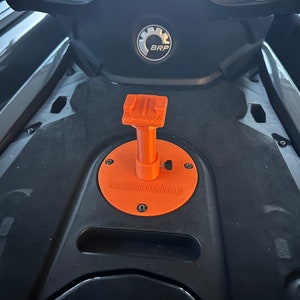 Puede incluir: Soporte de GoPro impreso en 3D de color naranja para una moto acuática. El soporte está fijado a la moto acuática con tornillos. El soporte tiene el texto "www.nrgcreativ.com" en él.