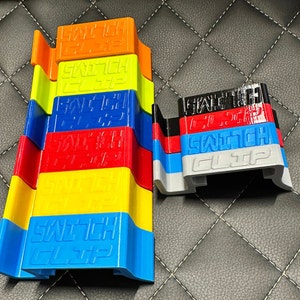 Könnte beinhalten: Ein Stapel aus sechs bunten Kunststoff-Schalterclips. Die Clips sind blau, gelb, rot, blau, gelb und orange. Der obere Clip trägt den Text "SWITCH CLIP" in schwarzen Buchstaben. Der untere Clip trägt den Text "SWITCH CLIP" in weißen Buchstaben.