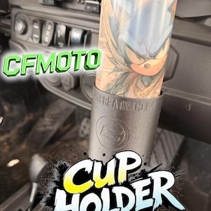 Puede incluir: Un portavasos negro con el texto "CUP HOLDER" en estilo cómic. El portavasos sostiene un vaso con un gráfico de Sonic the Hedgehog. También es visible el texto "CFMOTO".