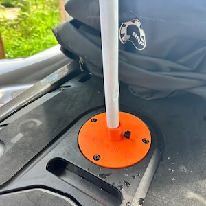 May include: A close-up of an orange flagpole mount with the text "NOW WE'RE CRAFTY.COM" on it. The mount is attached to a black surface and a white flagpole is inserted into the mount.
