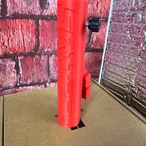 Puede incluir: Un soporte para tel&eacute;fono impreso en 3D de color rojo con la palabra "SWIMS" impresa en el lateral. El soporte tiene una perilla negra en la parte superior.