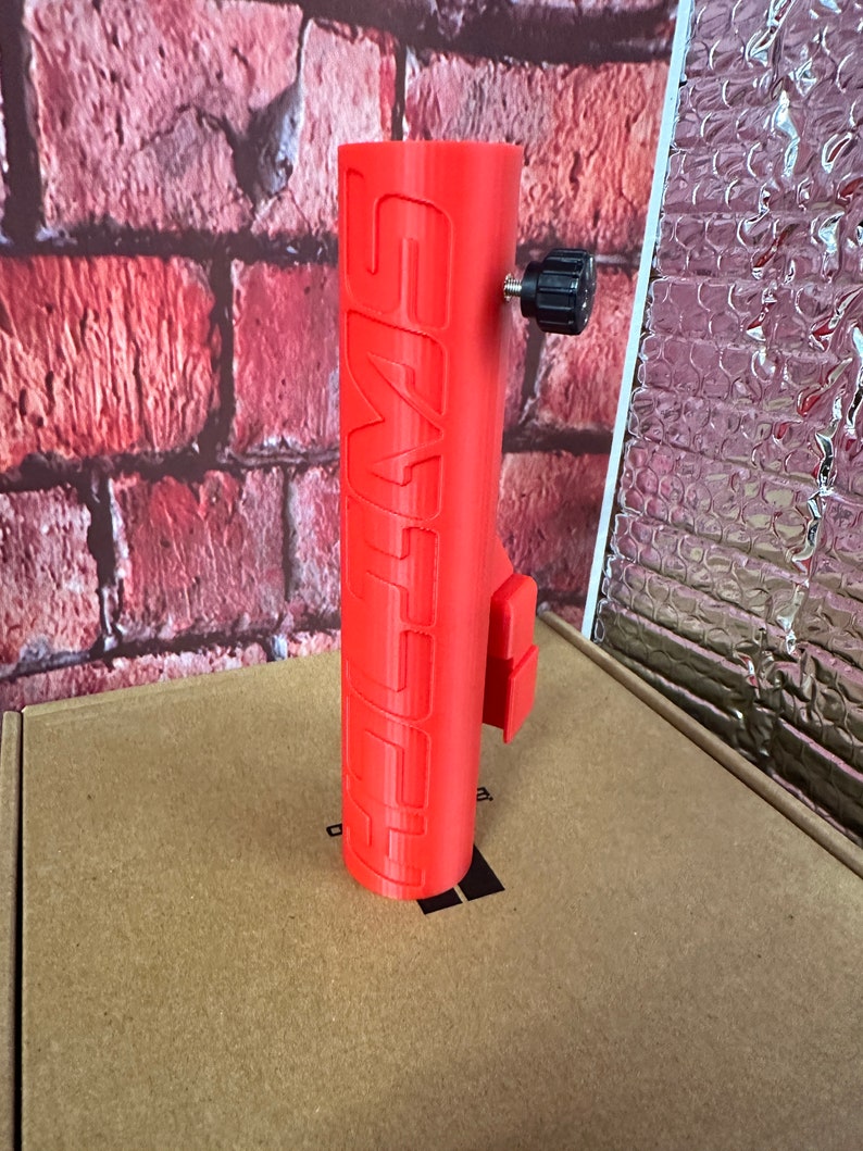 Puede incluir: Un soporte para tel&eacute;fono impreso en 3D de color rojo con la palabra "SWIMS" impresa en el lateral. El soporte tiene una perilla negra en la parte superior.