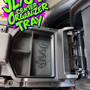 Op de afbeelding: Zwarte middenconsole organizer lade voor JL/JT Jeep modellen. De lade heeft drie compartimenten en is voorzien van het woord "Jeep" in reliëf op de bodem. De tekst "JL/JT CENTER ORGANIZER TRAY" wordt weergegeven in een kleurrijk graffiti-lettertype.