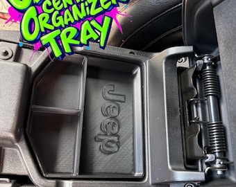 Könnte beinhalten: Schwarze Mittelkonsolen-Organizer-Ablage für JL/JT Jeep-Modelle. Das Tablett hat drei Fächer und trägt das Wort "Jeep" auf der Unterseite geprägt. Der Text "JL/JT CENTER ORGANIZER TRAY" wird in einer farbenfrohen Graffiti-Schriftart angezeigt.