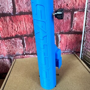 Puede incluir: Un soporte para tel&eacute;fono impreso en 3D de color azul con la palabra "SWIFT" impresa en el lateral. El soporte tiene una perilla negra para ajustar la tensi&oacute;n.