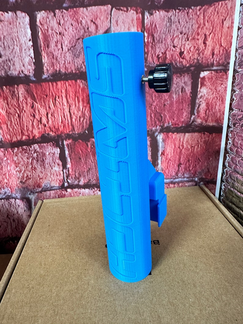 Puede incluir: Un soporte para tel&eacute;fono impreso en 3D de color azul con la palabra "SWIFT" impresa en el lateral. El soporte tiene una perilla negra para ajustar la tensi&oacute;n.