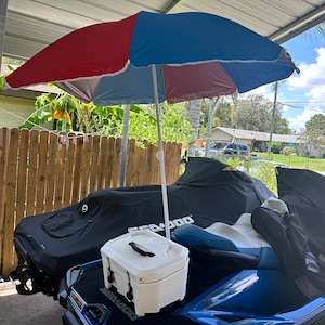 May include: A red, blue, and white beach umbrella is set up over two covered jet skis. A white cooler sits on the front of one jet ski. The jet skis are blue and black.