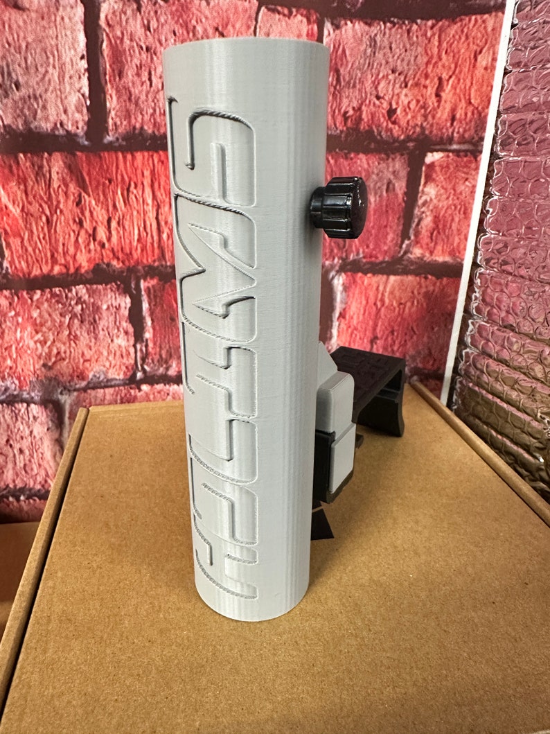 Puede incluir: Un soporte para tel&eacute;fono impreso en 3D de color gris con la palabra "SMYTH" impresa en el lateral. El soporte tiene una perilla negra para ajustar el &aacute;ngulo.