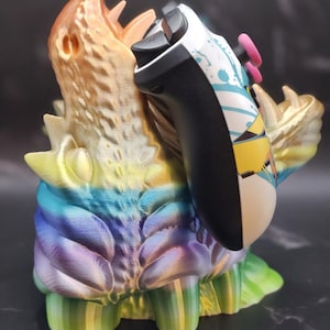 May include: A 3D printed, multi-colored dragon-like creature with a textured surface, holding a white and black video game controller with a blue, yellow, and white design.