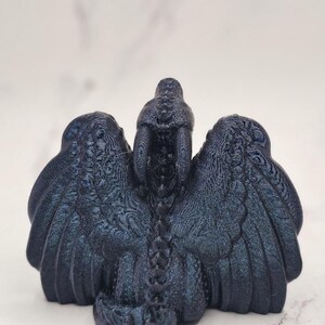 May include: A dark blue dragon figurine with outstretched wings. The sculpture has a textured surface and intricate details, including scales and feather-like patterns on the wings. The dragon is positioned against a light background.