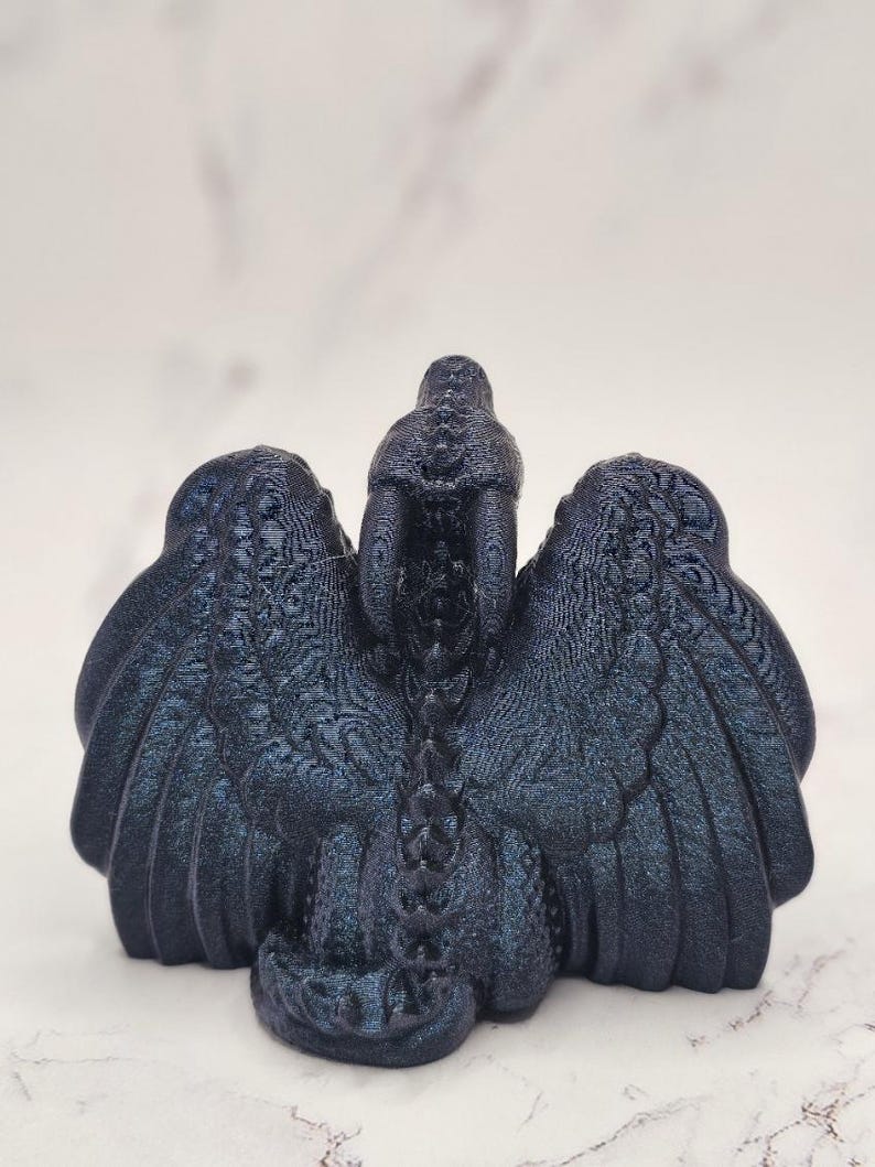 May include: A dark blue dragon figurine with outstretched wings. The sculpture has a textured surface and intricate details, including scales and feather-like patterns on the wings. The dragon is positioned against a light background.