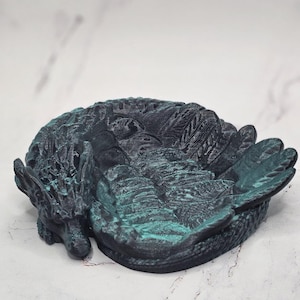 May include: A black and teal dragon sculpture curled up in a sleeping position. The dragon has detailed scales and wings.