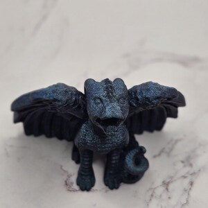 May include: A dark blue figurine of a mythical creature with outstretched wings, a reptilian body, and a curled tail. The sculpture has a textured surface and is set against a marble background. The creature's wings are spread wide, and its features are detailed.