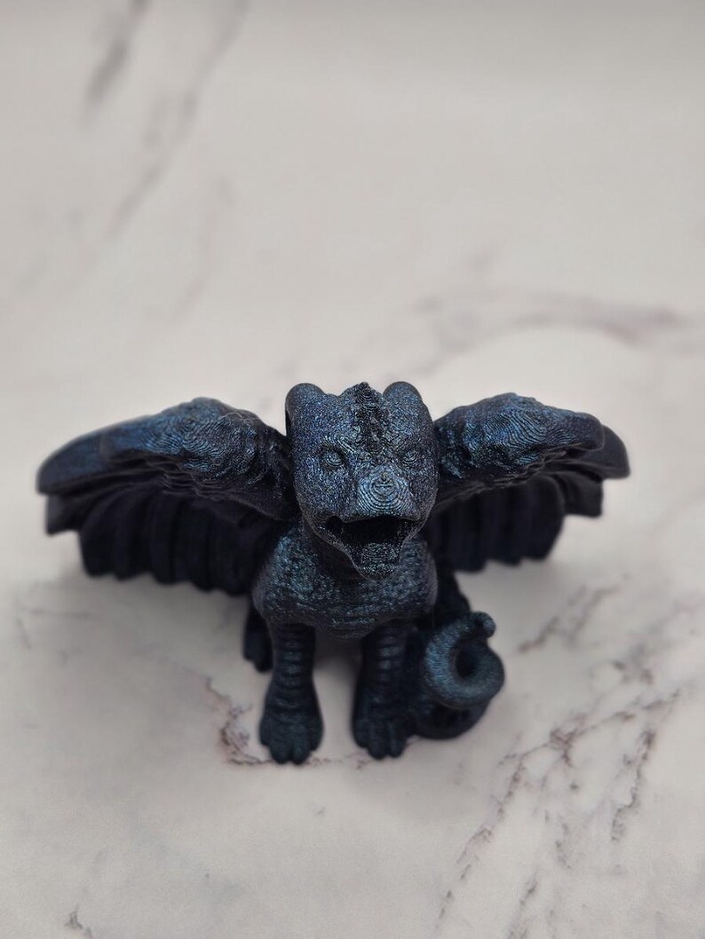 May include: A dark blue figurine of a mythical creature with outstretched wings, a reptilian body, and a curled tail. The sculpture has a textured surface and is set against a marble background. The creature's wings are spread wide, and its features are detailed.