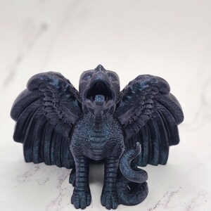 May include: A dark blue griffin figurine with outstretched wings and an open beak. The mythical creature has a textured surface and a curled tail. The figurine is standing on a white surface.